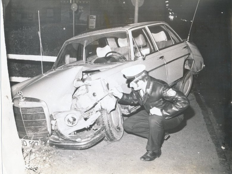 Black and white photograph Police officer kneels in front of demolished car