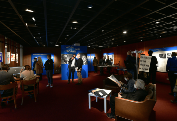 An unterschiedlichen Stationen schauen sich Besucherinnen und Besucher die Ausstellung an. Der Raum ist abgedunkelt. Das direkte Licht fällt vor allem auf die Ausstellungstafeln. 