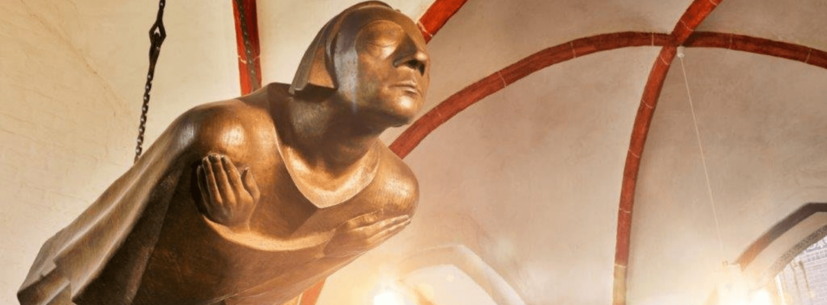 Eine Bronze-Skulptur eines Menschen hängt waagerecht von einem Kirchengewölbe. Die Hände sind überkreuz auf der Brust abgelegt, die Augen geschlossen.