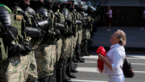 Demonstrator dressed in white with a red scarf in her hand kneels in front of a row of police officers in protective gear