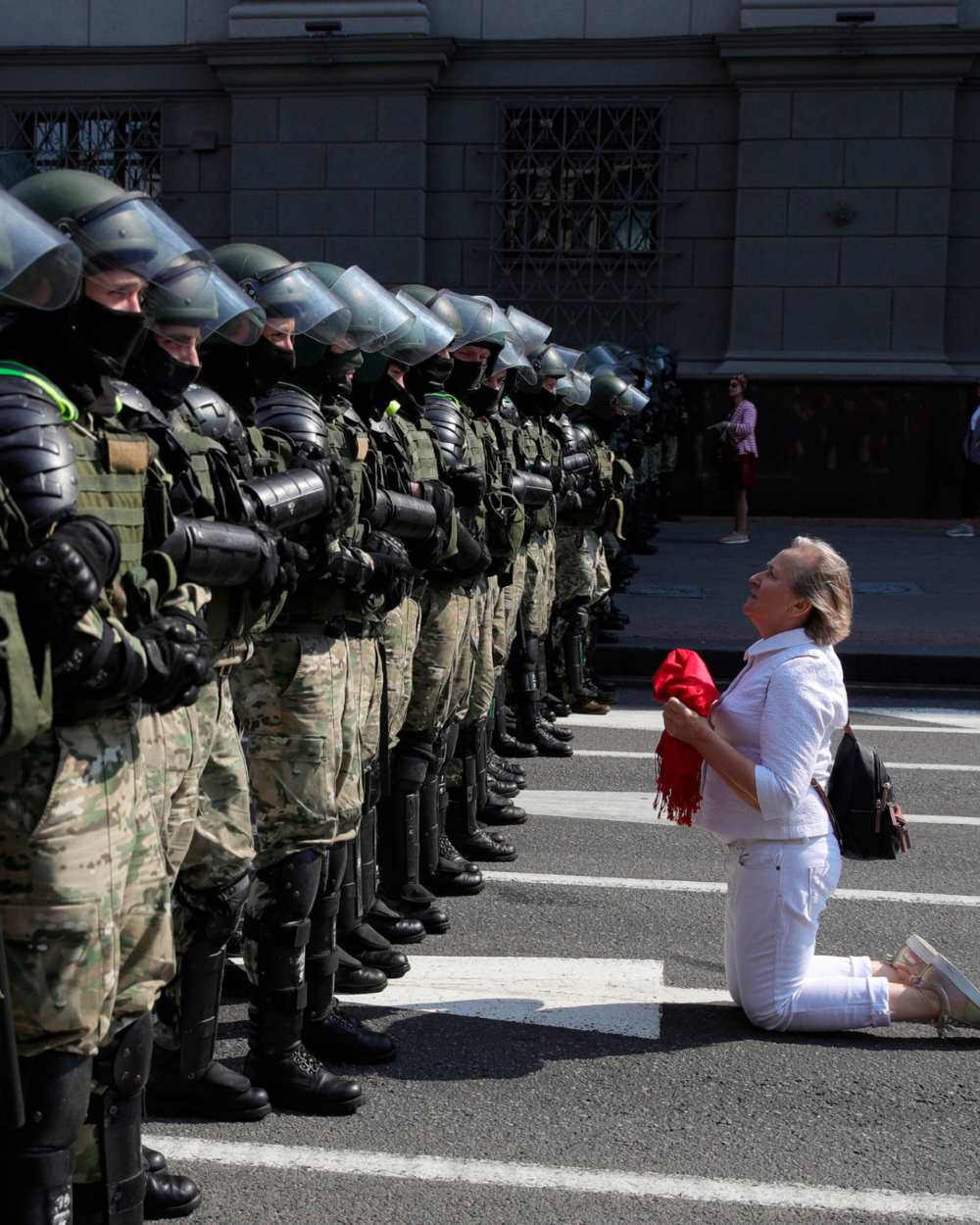 Demonstrator dressed in white with a red scarf in her hand kneels in front of a row of police officers