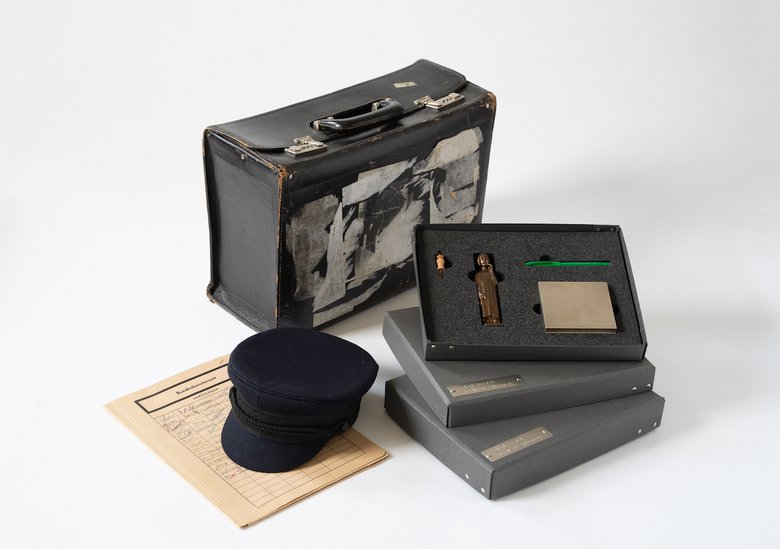 colour photo: exhibition items you can touch. A cap lies on a sheet of paper, with two cardboard boxes filled with foam and objects to the right of it. A suitcase can be seen in the background.