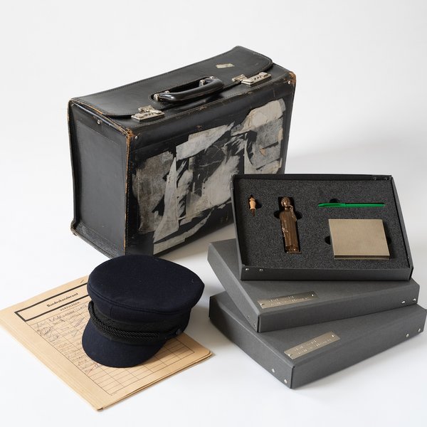 colour photo: exhibition items you can touch. A cap lies on a sheet of paper, with two cardboard boxes filled with foam and objects to the right of it. A suitcase can be seen in the background.