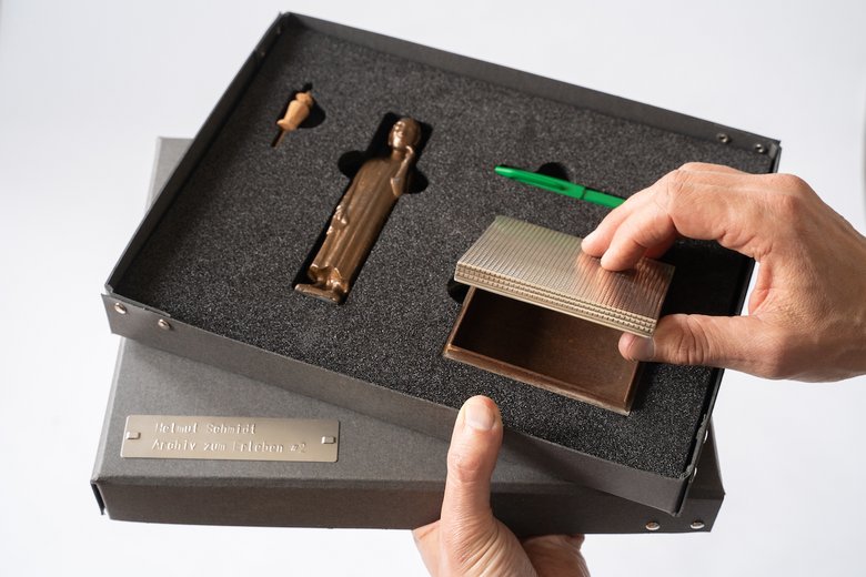 Colour photo: an open box containing exhibition items that visitors can touch. On the left, a pin and a small bronze statue. On the right, a small tin and a green pen.
