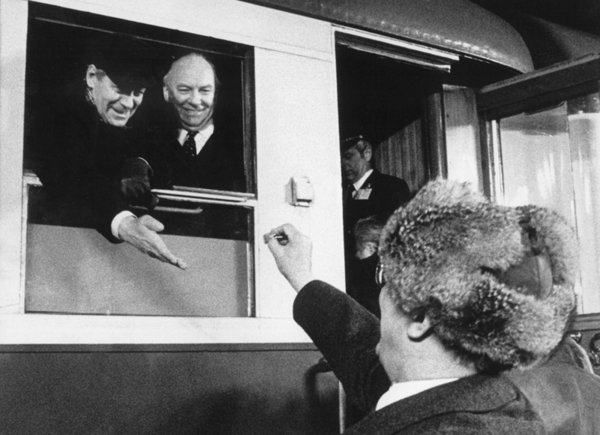 Helmut Schmidt streckt seine Hand aus einem geöfneten Fenster eines Zuges, während Erich Honecker ihm vom Bahngleis ein Bonbon in die Hand legt.