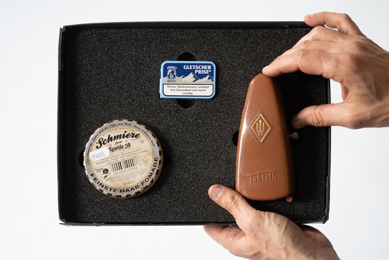Colour photo: An open cardboard box containing foam. The foam holds exhibition items that visitors can touch: a hair pomade, a tin of snuff and a Taktell.