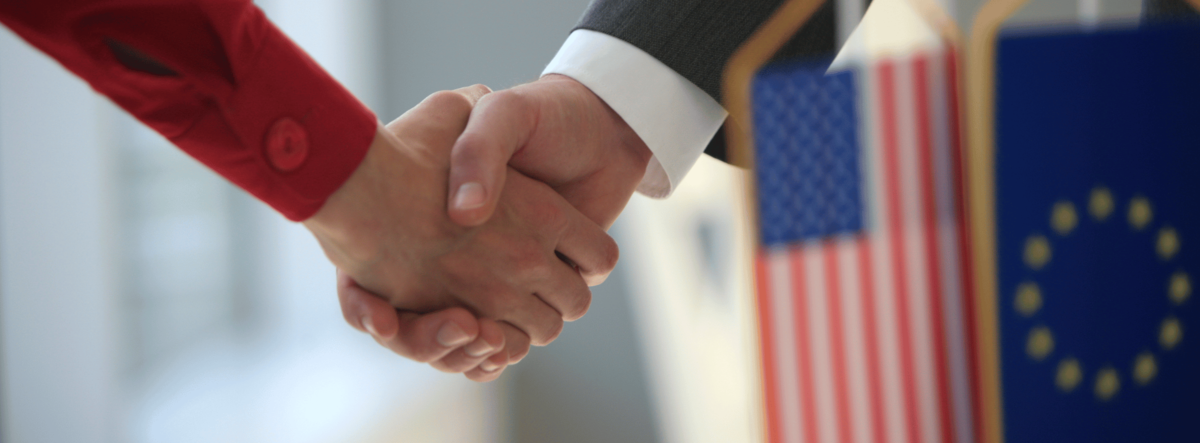 Nahaufnahme eines Händedrucks zwischen zwei Personen neben einer US-amerikanischer Flagge und europäischer Flagge.