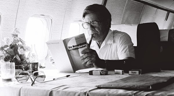 Während eines Fluges liest Helmut Schmidt das Buch „Die Neuordnung der Weltwirtschaft“. Vor ihm stehen eine Cola-Flasche und einige Zigarettenschachteln.