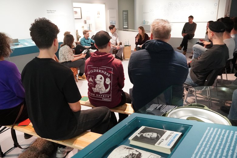 Menschen sitzen beieinander und sprechen. Daneben Ausstellungselemente zum Thema Demokratie.