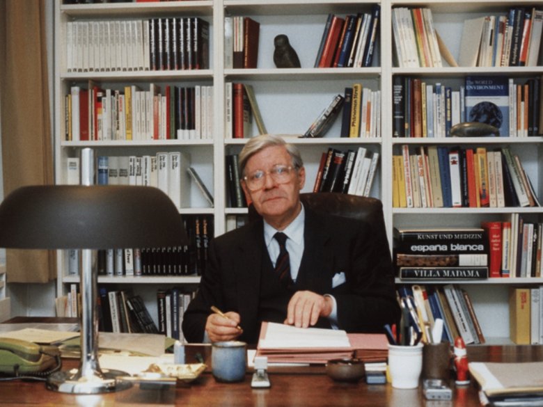 Helmut Schmidt sitting at a desk in front of a wall cupboard with books