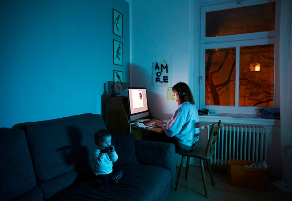 Frau arbeitet abends am Computer, neben ihr sitzt ein Kleinkind auf einer Couch.