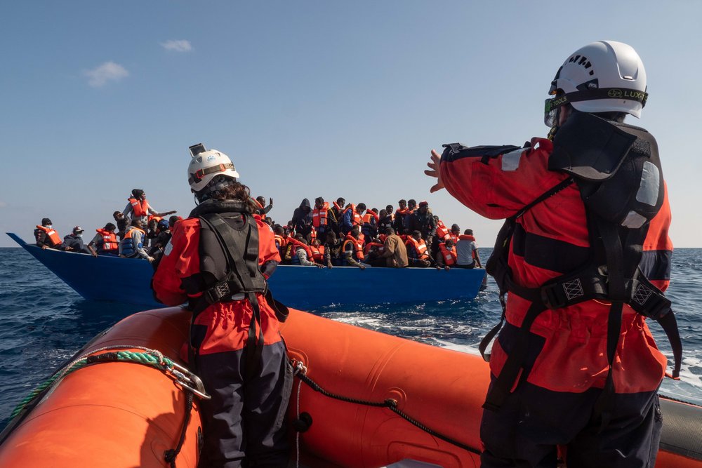Rotes Boot der Sea-Watch mit zwei Personen vor blauem Boot mit zahlreichen Menschen auf Meer