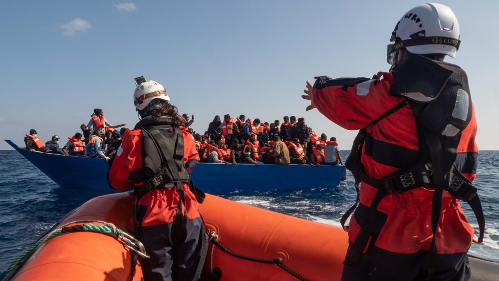 Rotes Boot der Sea-Watch mit zwei Personen vor blauem Boot mit zahlreichen Menschen auf Meer