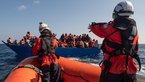 Rotes Boot der Sea-Watch mit zwei Personen vor blauem Boot mit zahlreichen Menschen auf Meer