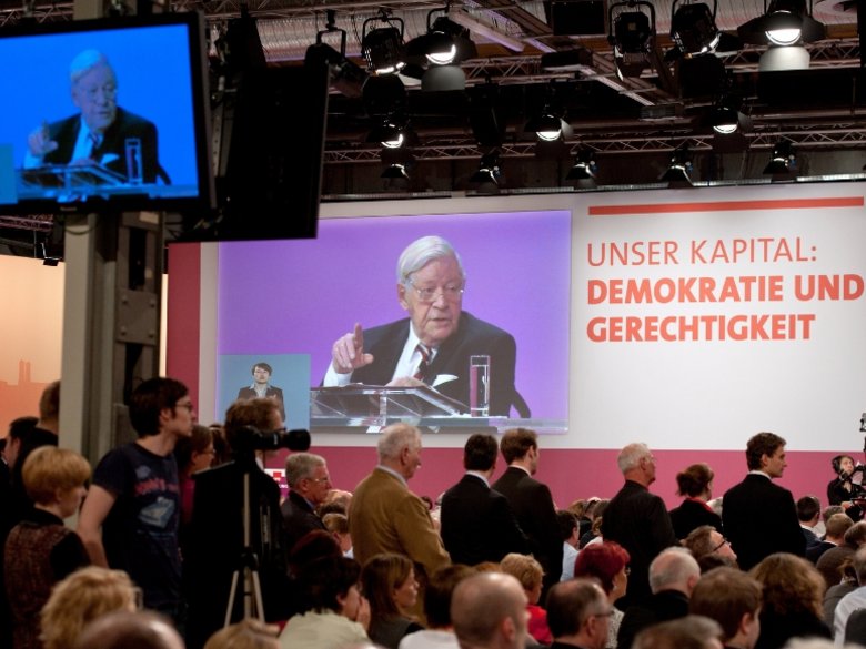 Helmut Schmidt gives a speech in 2011 and is projected onto the wall. An audience sits in front of him