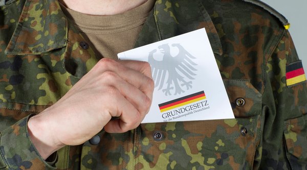 Moderne Aufnahme zeigt einen Mann in Bundeswehrbekleidung. Er ist im Begriff eine Ausgabe des deutschen Grundgesetzes in seiner Brusttasche zu verstauen.