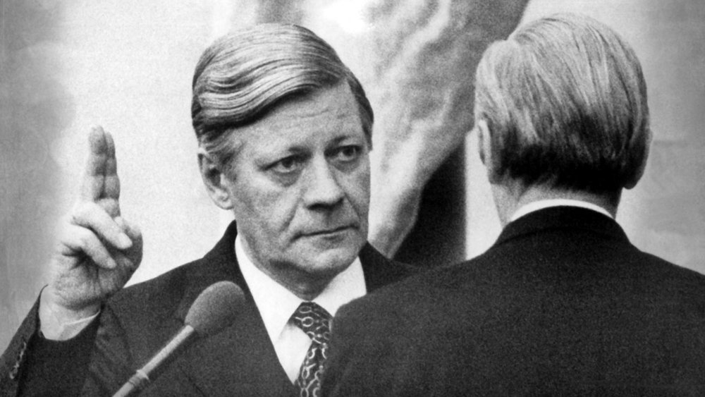 Schwarzweißfoto: Helmut Schmidt hebt die rechte Hand und blickt ernst zu einem Mann, der ihm gegenübersteht; ein Mikrofon ragt ins Bild. Knapp zwei Stunden nach seiner erneuten Wahl durch den Deutschen Bundestag wird Helmut Schmidt (SPD) am 15. Dezember 1976 von Parlamentspräsident Karl Carstens als Bundeskanzler vereidigt. 