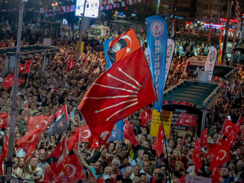 A crowd of people at a political rally with many Turkish flags and red party flags waving above the crowd