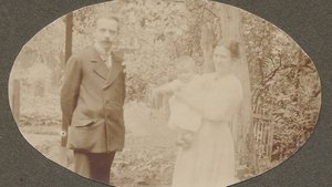 Historical sepia photograph from 1919: Helmut Schmidt as a toddler in his mother’s arms, with his father in a suit standing beside them; taken outdoors in front of trees and a garden fence.