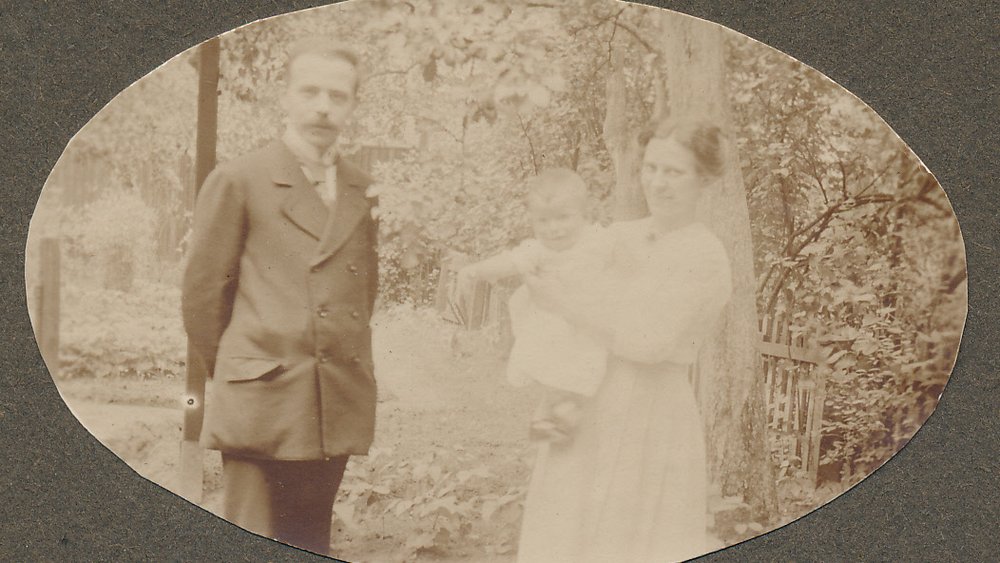 Historical sepia photograph from 1919: Helmut Schmidt as a toddler in his mother’s arms, with his father in a suit standing beside them; taken outdoors in front of trees and a garden fence.