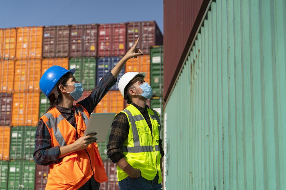 Workers with helmets and high-visibility waistcoats in front of harbour containers