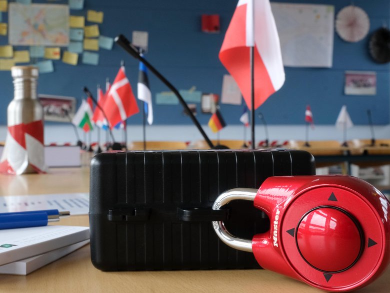 Colour photo: The setup for the escape game. A locked suitcase on a table, with flags of European countries in the background