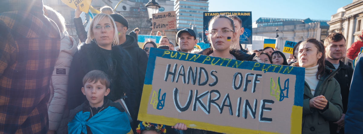 Demonstrierende, darunter auch Kinder, mit Protestschildern. Eines sagt „Hands off Ukraine“.