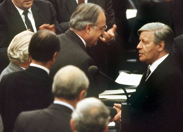 Helmut Kohl und Helmut Schmidt geben sich die Hand im Bonner Bundestag. Um sie herum sind weitere Bundestagsabgeordnete.