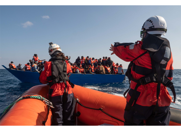 Rettungspersonen in einem Schlauchboot steuern blaues Boot zu, das mit Menschen mit Schwimmwesten überfüllt ist.