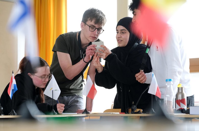 Colour photo: Blurred flags in the foreground. In the background, a group of young people sitting at a table. They are chatting whilst playing the escape game Unlock Europe.