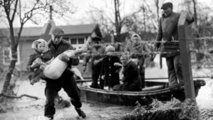 Black-and-white photograph of the 1962 storm surge: a man carries a woman through the water; behind him is a boat carrying several evacuees. During the major storm surge disaster on the night of 16–17 February 1962, 315 people lost their lives, 200 of them in the Wilhelmsburg district of Hamburg alone.