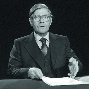 Black-and-white photograph, 1977: Helmut Schmidt sitting at a table during a televised statement regarding the kidnapping of Hanns Martin Schleyer, President of the Confederation of German Employers’ Associations.. He is wearing glasses and a suit, with a pile of papers in front of him.