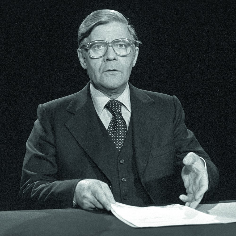 Black-and-white photograph, 1977: Helmut Schmidt sitting at a table during a televised statement regarding the kidnapping of Hanns Martin Schleyer, President of the Confederation of German Employers’ Associations.. He is wearing glasses and a suit, with a pile of papers in front of him.