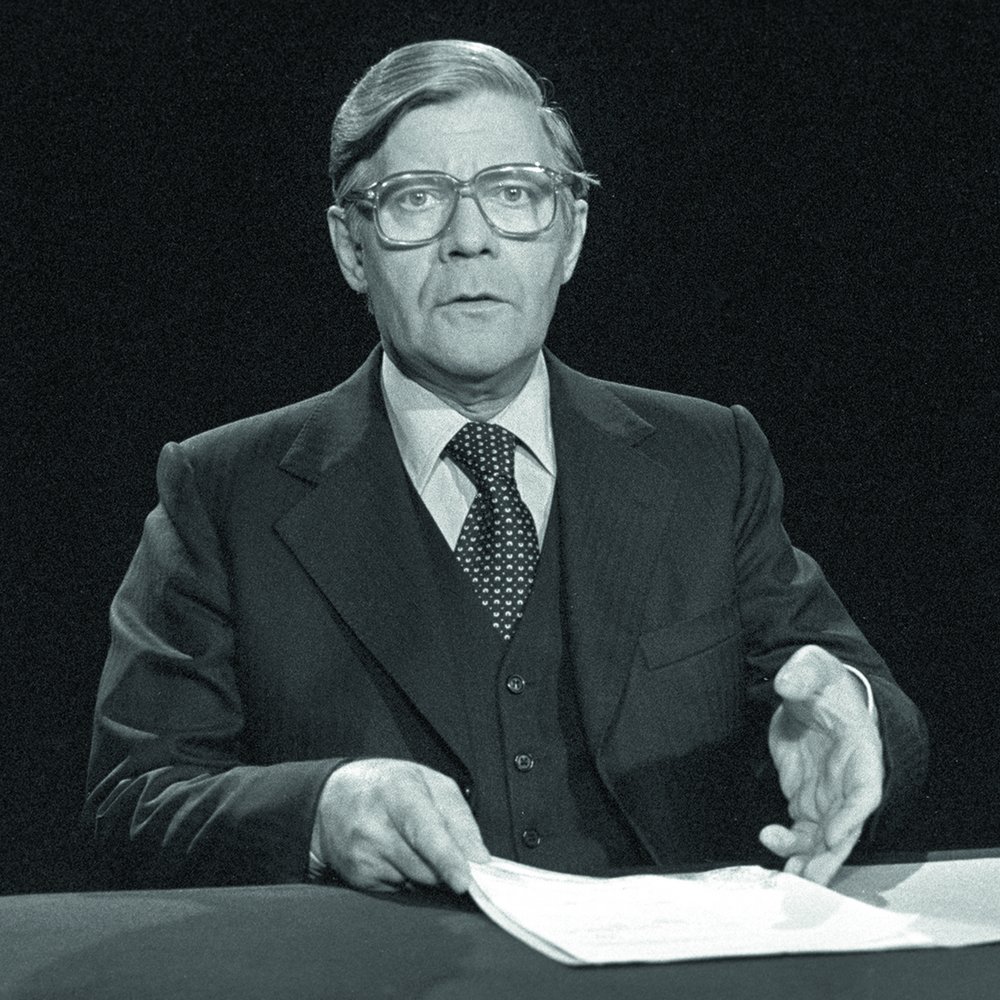 Schwarzweißfoto 1977: Helmut Schmidt in einer Fernsehansprache am Tisch, mit Brille und Anzug, vor ihm ein Stapel Papiere.