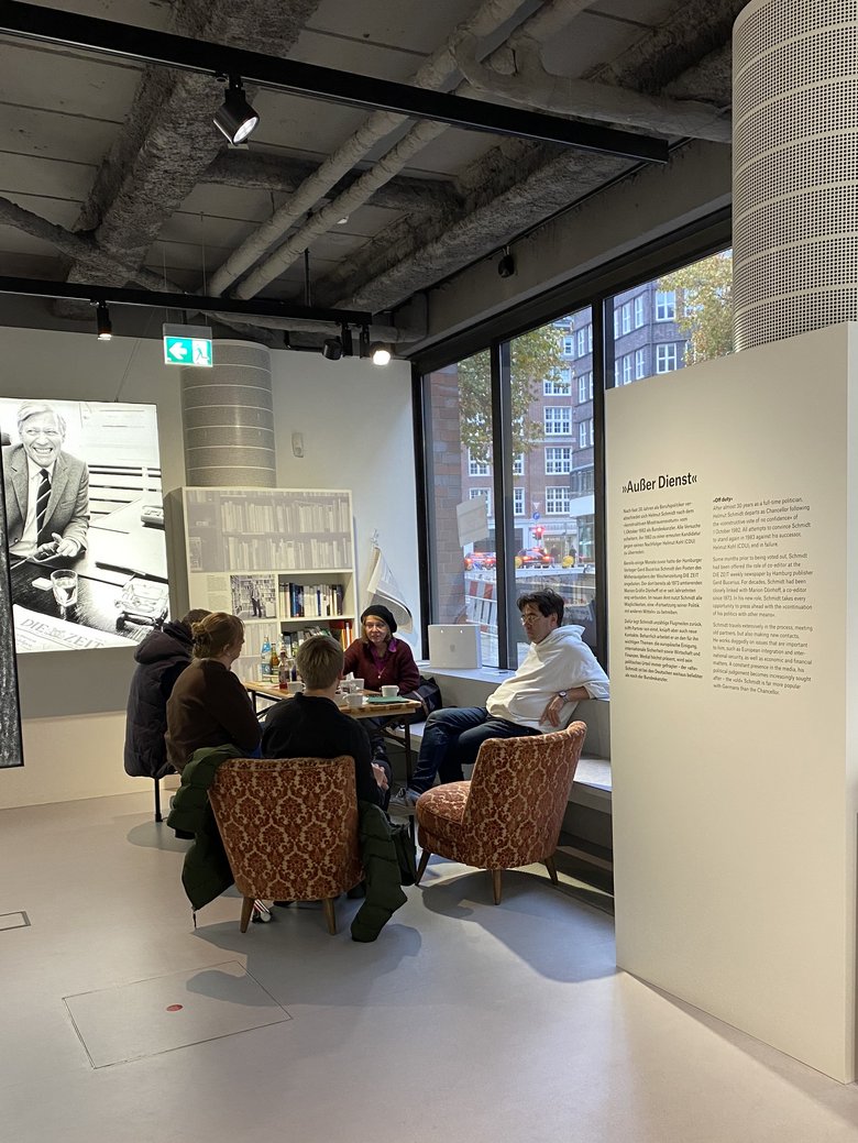 People sit in a circle and talk. Next to them are exhibition elements on Helmut Schmidt.