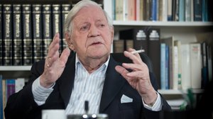 Colour photograph: Helmut Schmidt in his office in front of bookshelves, explaining something with his hands raised; a cigarette between his fingers.