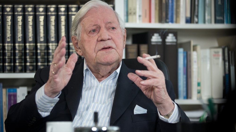 Colour photograph: Helmut Schmidt in his office in front of bookshelves, explaining something with his hands raised; a cigarette between his fingers.