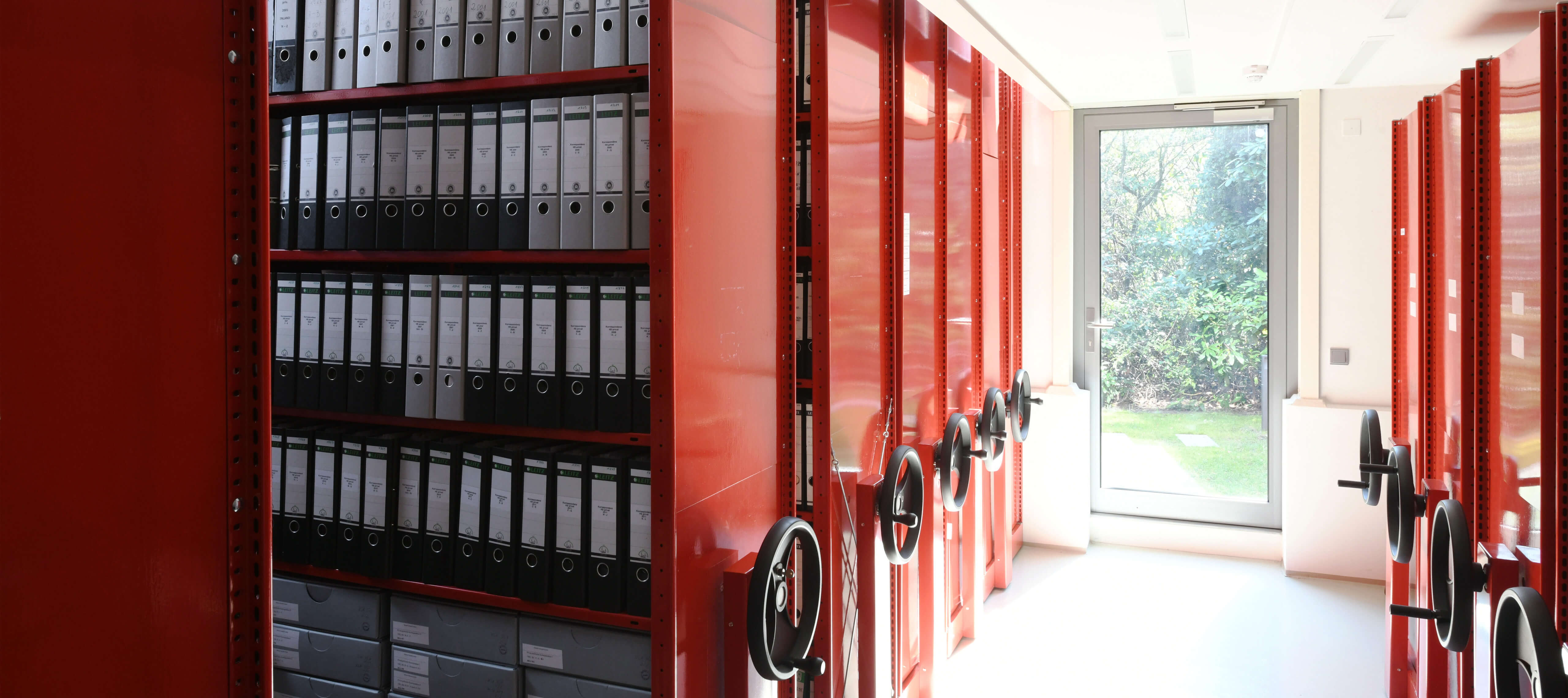 Blick in das Archiv. Die Aktenordner werden in roten bewegbaren Archiv-Regalen aus Blech aufbewahrt.