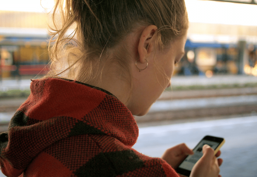  Rückenansicht einer jungen Frau, die auf ein Smartphone schaut.