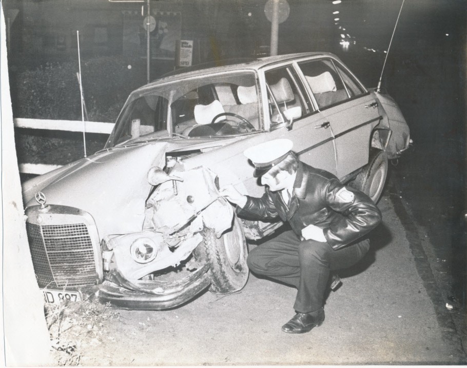 Black and white photograph of kneeling policeman in front of demolished car