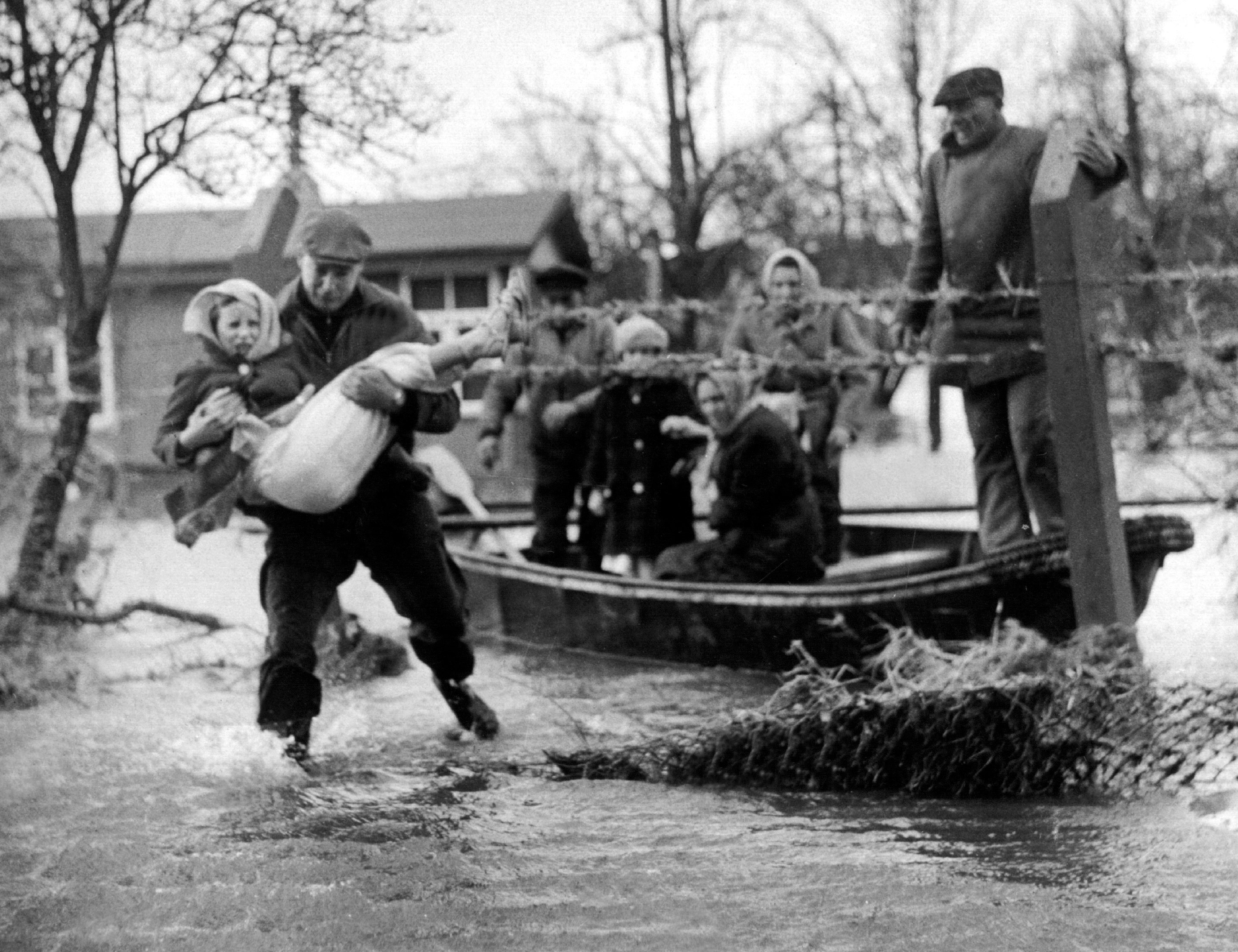 Schwarzweißfoto der Sturmflut 1962: Ein Mann trägt eine Frau durch das Wasser; hinter ihm ein Boot mit mehreren Evakuierten. Bei der großen Sturmflutkatastrophe in der Nacht vom 16. auf den 17.02.1962 starben 315 Menschen, davon alleine 200 in dem Hamburger Stadtteil Wilhelmsburg.
